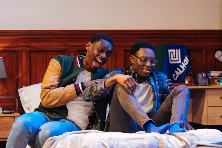 Two young men are sat on a bed. One is laughing hysterically and grabbing the other's arms, while the other smiles awkwardly.
