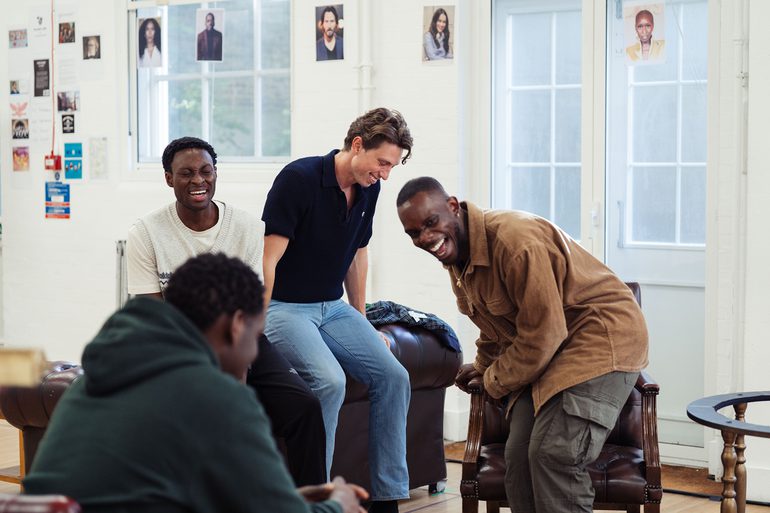 Four young men laugh together. Two sit on the back of an old-fashioned leather sofa, one stands bent over with laughter, and the other has his back to the camera.