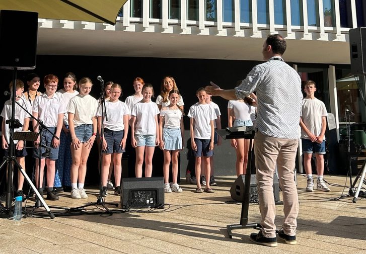 A group of young people are stood outside with their arms by their sides. They are all wearing white tops and blue denim shorts. There are large speakers either side of the group and some microphones in front of them. To the left, there is someone sat at a keyboard with sheet music in front of them. Facing the group is a conductor who is wearing a white patterned shirt and beige trousers. He has a music stand next to him and has his arms extended in front of him.