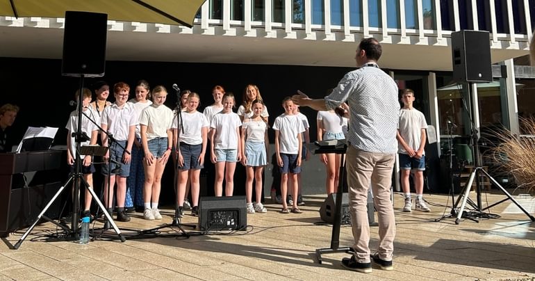 A group of young people are stood outside with their arms by their sides. They are all wearing white tops and blue denim shorts. There are large speakers either side of the group and some microphones in front of them. To the left, there is someone sat at a keyboard with sheet music in front of them. Facing the group is a conductor who is wearing a white patterned shirt and beige trousers. He has a music stand next to him and has his arms extended in front of him.