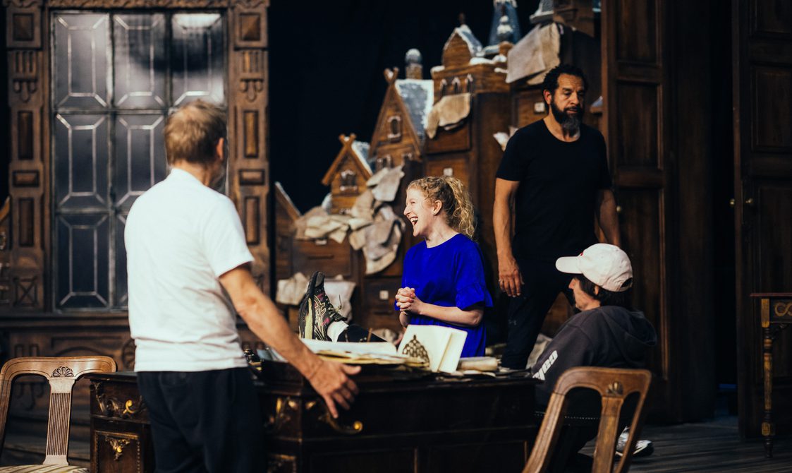 A group of actors are gathered around the set during warm-up, the men have their backs to the camera but a woman with curly hair is smiling, verging on laughter.