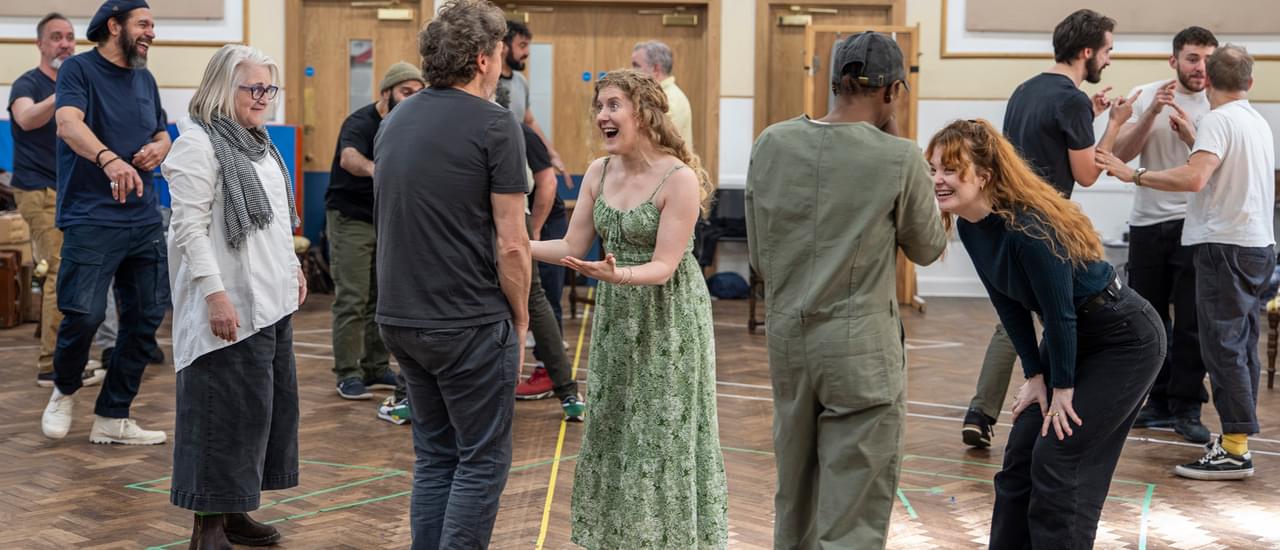 Performers wearing casual clothes rehearse in a studio. The room is crowded with smaller groups forming, talking and laughing together. In the centre, four performers stand facing one another in two lines. The two women facing the camera are laughing and smiling with animated expressions.