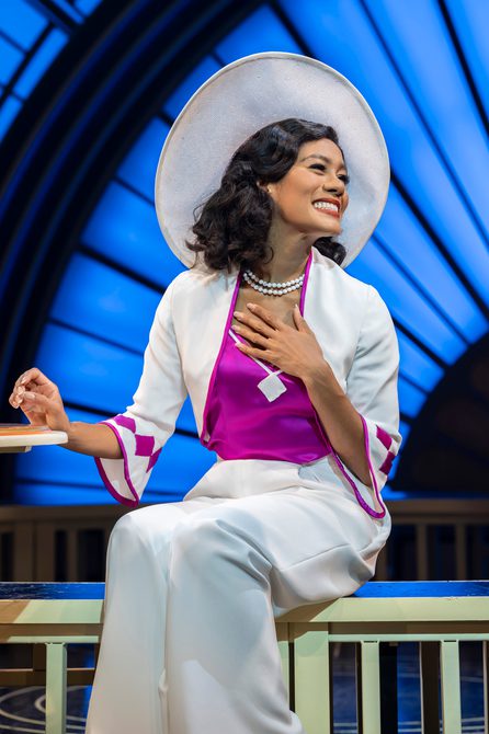 A woman is sat elegantly with one hand on her chest and the other resting on the table next to her. She is wearing a large white hat, a white satin bolero jacket with fuschia piping and diamond details on the cuffs. Her top is in the same fuschia and her trousers match the hat and jacket. She has two simple strings of pearls. She is smiling brightly.