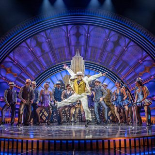 A company of dancers are all wearing refined 1930s outfits. The men have cream fedoras, matching waistcoats and trousers, striped shirts and polka ties. The women have mini A-line dresses and fascinators with striped bows. Front and centre, a man in a cream suit and matching hat with a mustard waistcoats dancing with his arms above his head and has legs kicking. The set is a detailed art deco arch with skyscrapers in front.