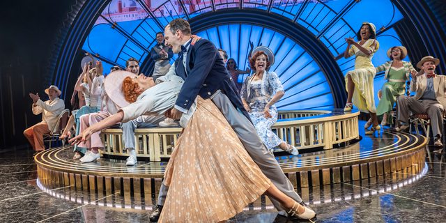 A man and woman are dancing. He is holding her by her waist as he dips her. She is leaning deeply with one arm reaching behind her and the other grasping his neck. He has on an sophisticated navy and grey tailed suit. She is wearing a light orange pleated skirt with white floral details, a white cropped jacket and large white hat. There are many performers behind them clapping and cheering. The backdrop is a large art deco arch with a projection of Venice.