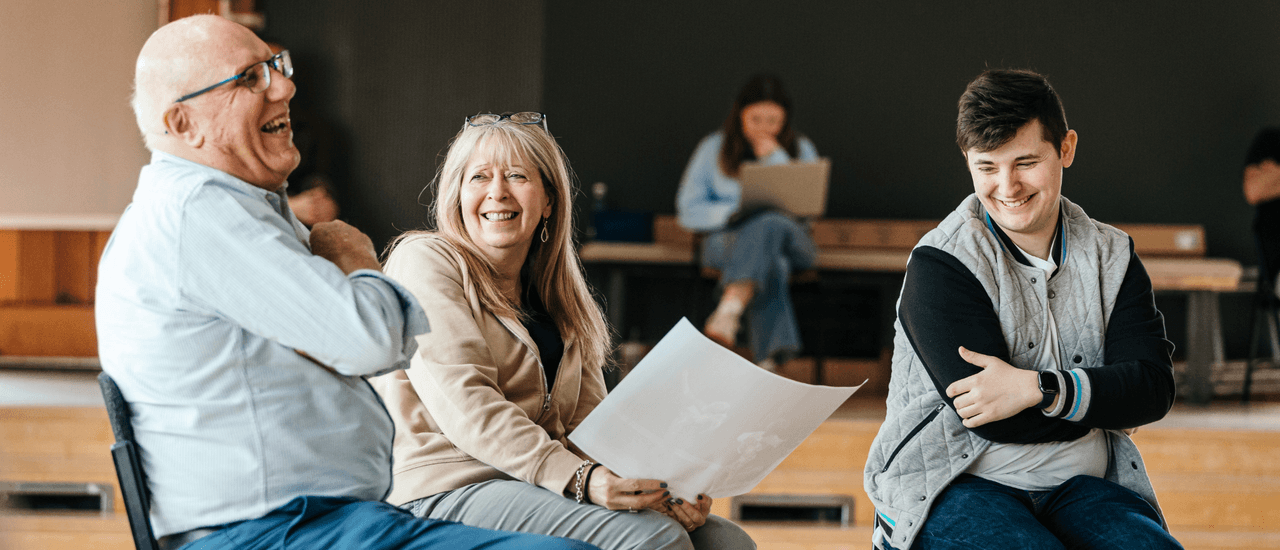 A man with white hair and glasses laughs while sat next to a smiling blonde woman (holding a large white piece of paper)and younger man with black hair.