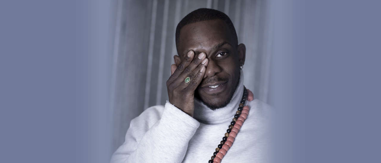 A headshot of director Roy Alexander Weise holding a hand over one eye and looking at the camera with a slight smile. He wears a light grey long sleeve turtle neck top and two chunky, long, wooden beaded necklaces.
