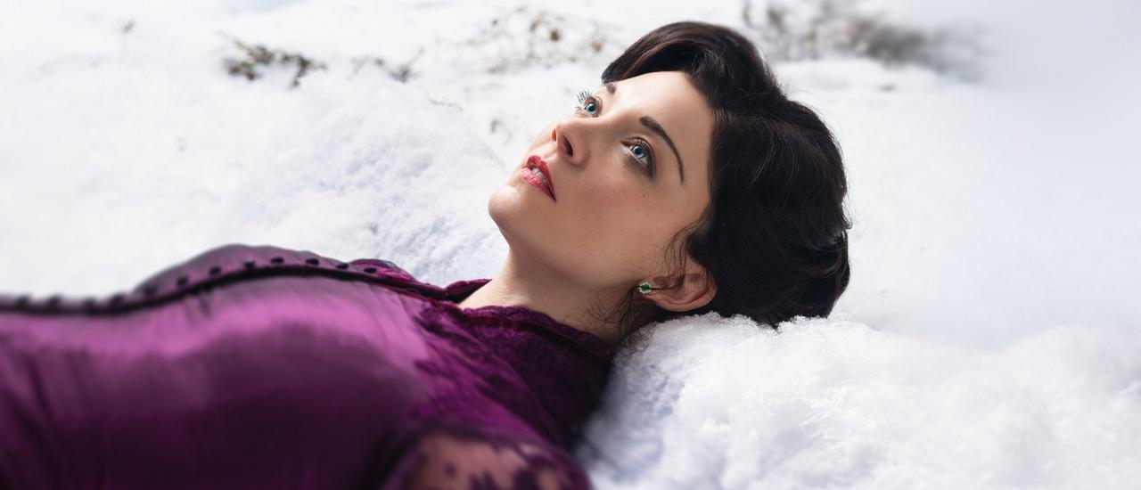 A young woman lays in the snow, looking up to the sky. She wears a rich purple silk dress with a high collar. She has brown hair and striking blue eyes. Her expression is numb and dissociated.