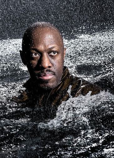 A man's head and shoulders rise from a sea of rough, churning water; rain is pouring down on him. He stares into the distance, his face illuminated by a shaft of light. He has dark hair and a moustache.