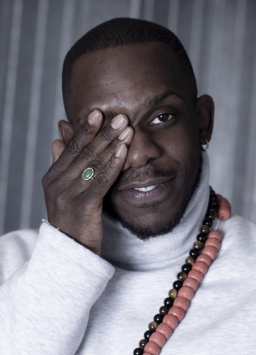 A headshot of director Roy Alexander Weise holding a hand over one eye and looking at the camera with a slight smile. He wears a light grey long sleeve turtle neck top and two chunky, long, wooden beaded necklaces.