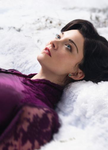 A young woman lays in the snow, looking up to the sky. She wears a rich purple silk dress with a high collar. She has brown hair and striking blue eyes. Her expression is numb and dissociated.