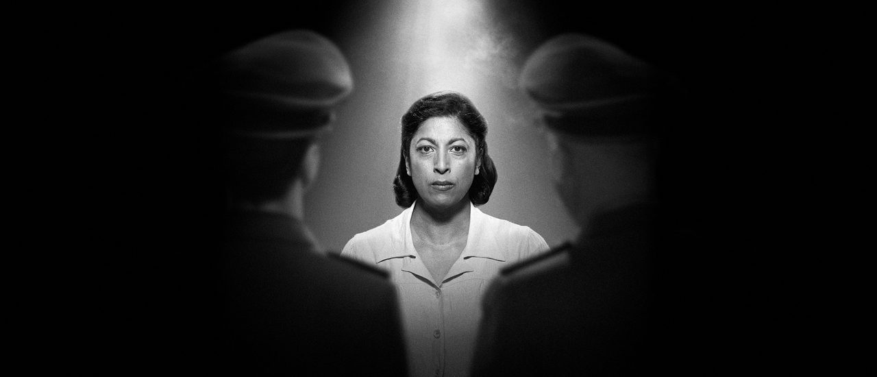 In a black and white photograph, a woman with dark shoulder-length hair stares resolutely at the camera, wearing a white top. A light illuminates her from above. Slightly out of focus, facing her on either side, are two men in military uniform with their backs to the camera.