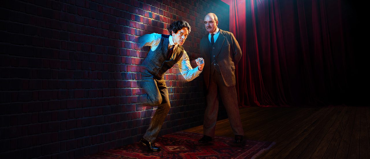 A dark-haired man with a determined expression is running through a brick wall. A stark spotlight is shining directly on him. A man with a neat moustache is looking at him intently with his hands behind his back.