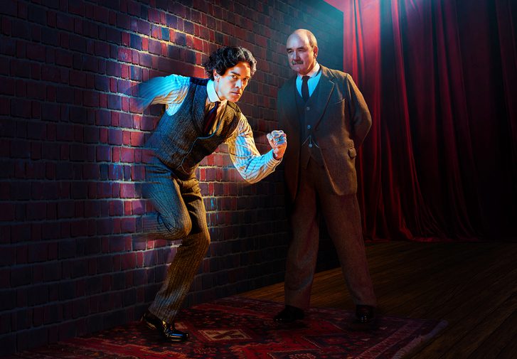 A dark-haired man with a determined expression is running through a brick wall. A stark spotlight is shining directly on him. A man with a neat moustache is looking at him intently with his hands behind his back.