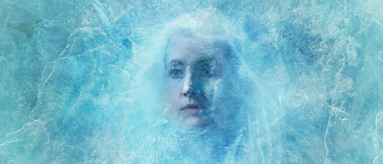 A woman frozen behind a sheet of ice - her hair is pulled out of her face to show her expression, also frozen in time.