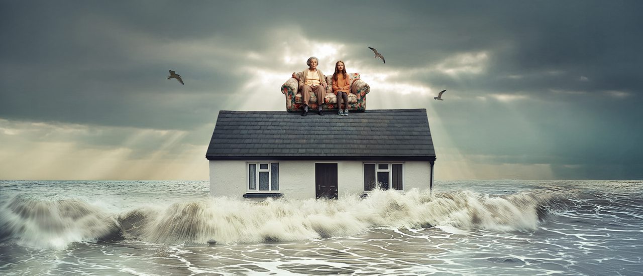 Grey ocean waves crash against the sides of a home. On the roof of the house, is a floral sofa with a young girl and her grandmother sat on, they both look concerned. The sky behind them is grey and cloudy, and a couple of seagulls fly around them.