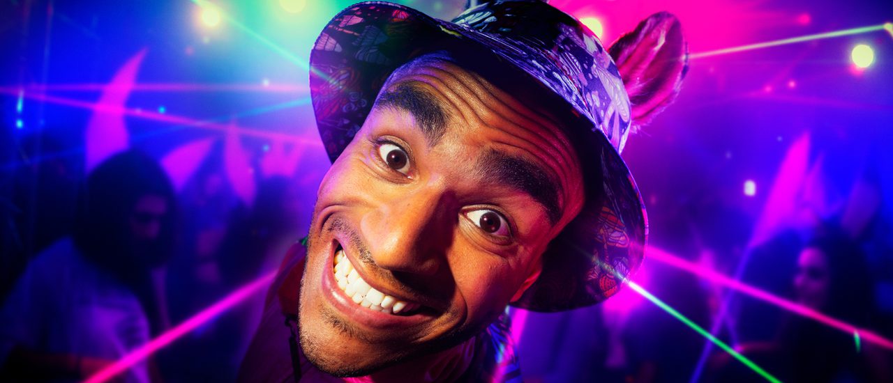 A young man is grinning intensely at the camera. He is wearing a bucket hat with donkey ears. The background is a colourful rave with purple and green strobe lights.