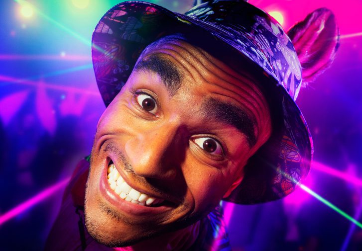 A young man is grinning intensely at the camera. He is wearing a bucket hat with donkey ears. The background is a colourful rave with purple and green strobe lights.