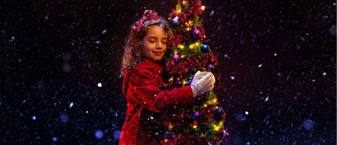A young girl in a red winter coat hugs a small Christmas tree. She is smiling contentedly with her eyes closed, and has a red tartan bow in her hair. The tree is decorated with red tinsel, purple baubles and gold twinkly lights. Snow is falling around them.
