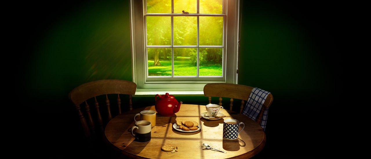 Three mugs of tea, a cup and saucer, a red teapot and a plate of digestive biscuits sit on a circular pine table. One slightly crumbled biscuit lies on its own, next to a used teaspoon. Sunlight floods in through the window behind, looking onto lush green grass and trees.