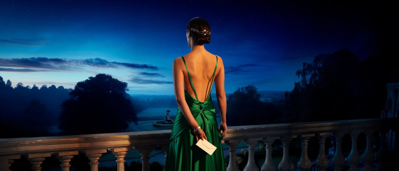 A young woman in a striking green backless dress holds a white envelope in one hand behind her back. She looks out at a garden and fountain from a balcony at dusk.