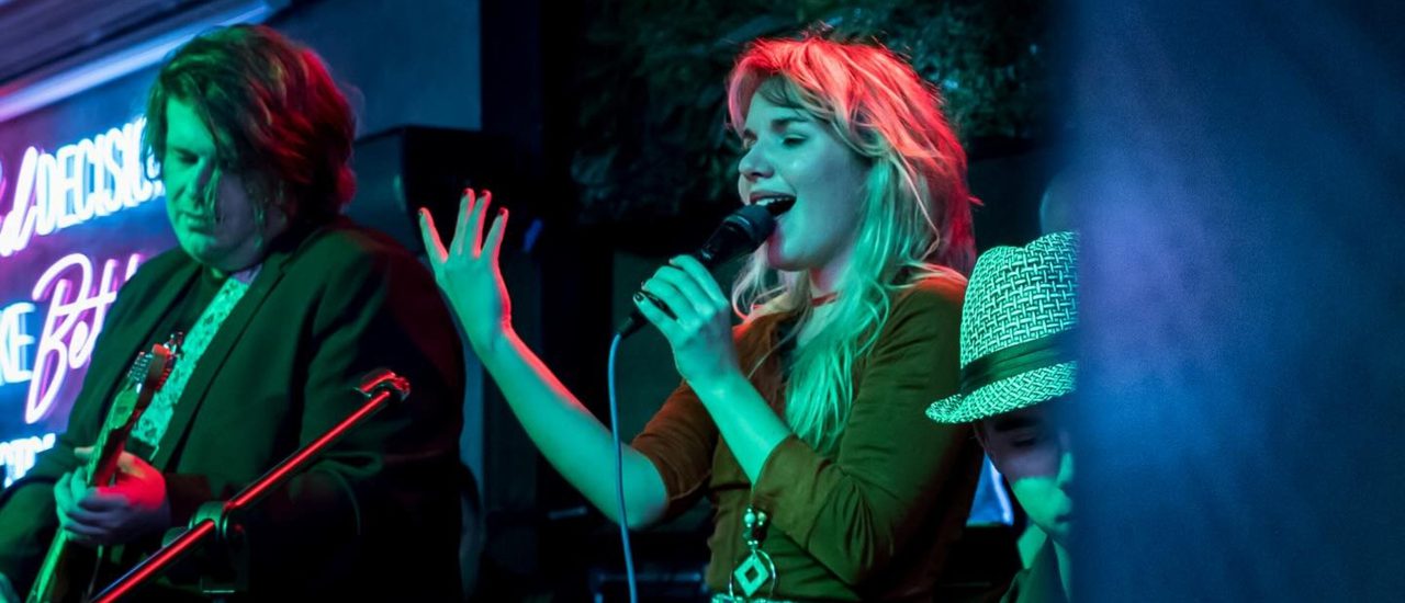 A band performing on stage under blue lighting, featuring a blonde female singer in the foreground, with a neon sign glowing behind them.