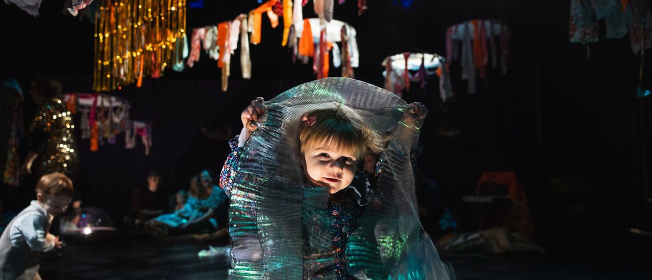 A small child hides in an iridescent fabric; colourful garlands and bunting are hanging in the background.