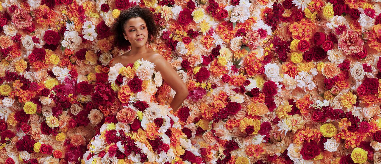 A young woman stands totally surrounded by flowers. They flood the background and cover her strapless, floor-length gown. The flowers are a mixture of pink, white, yellow and orange tones. The woman holds her hand behind her back and looks off to the side with a content, quietly confident facial expression.
