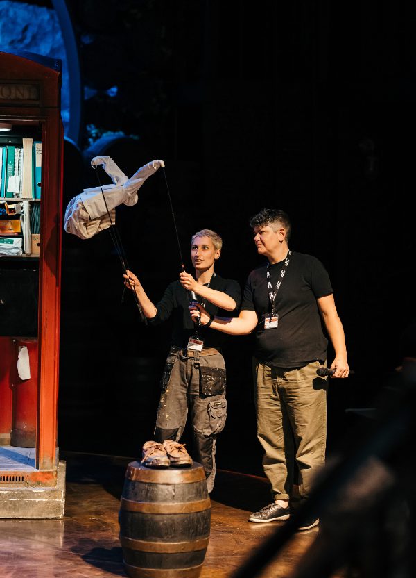 Two prop makers in black t-shirts and beige trousers are operating a small cloth puppet on stage.