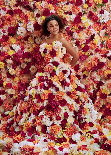 A young woman stands totally surrounded by flowers. They flood the background and cover her strapless, floor-length gown. The flowers are a mixture of pink, white, yellow and orange tones. The woman holds her hand behind her back and looks off to the side with a content, quietly confident facial expression.