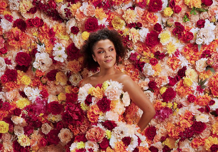 A young woman stands totally surrounded by flowers. They flood the background and cover her strapless, floor-length gown. The flowers are a mixture of pink, white, yellow and orange tones. The woman holds her hand behind her back and looks off to the side with a content, quietly confident facial expression.