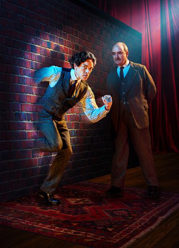 A dark-haired man with a determined expression is running through a brick wall. A stark spotlight is shining directly on him. A man with a neat moustache is looking at him intently with his hands behind his back.