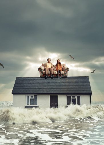 Grey ocean waves crash against the sides of a home. On the roof of the house, is a floral sofa with a young girl and her grandmother sat on, they both look concerned. The sky behind them is grey and cloudy, and a couple of seagulls fly around them.