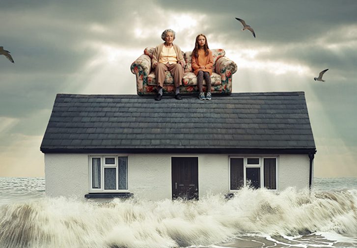 Grey ocean waves crash against the sides of a home. On the roof of the house, is a floral sofa with a young girl and her grandmother sat on, they both look concerned. The sky behind them is grey and cloudy, and a couple of seagulls fly around them.
