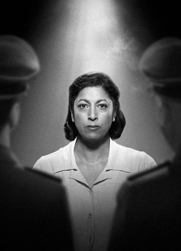 In a black and white photograph, a woman with dark shoulder-length hair stares resolutely at the camera, wearing a white top. A light illuminates her from above. Slightly out of focus, facing her on either side, are two men in military uniform with their backs to the camera.