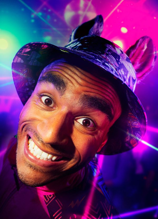 A young man is grinning intensely at the camera. He is wearing a bucket hat with donkey ears. The background is a colourful rave with purple and green strobe lights.
