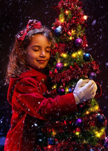 A young girl in a red winter coat hugs a small Christmas tree. She is smiling contentedly with her eyes closed, and has a red tartan bow in her hair. The tree is decorated with red tinsel, purple baubles and gold twinkly lights. Snow is falling around them.