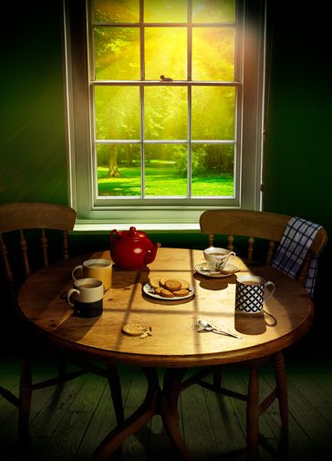 Three mugs of tea, a cup and saucer, a red teapot and a plate of digestive biscuits sit on a circular pine table. One slightly crumbled biscuit lies on its own, next to a used teaspoon. Sunlight floods in through the window behind, looking onto lush green grass and trees.
