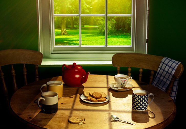 Three mugs of tea, a cup and saucer, a red teapot and a plate of digestive biscuits sit on a circular pine table. One slightly crumbled biscuit lies on its own, next to a used teaspoon. Sunlight floods in through the window behind, looking onto lush green grass and trees.