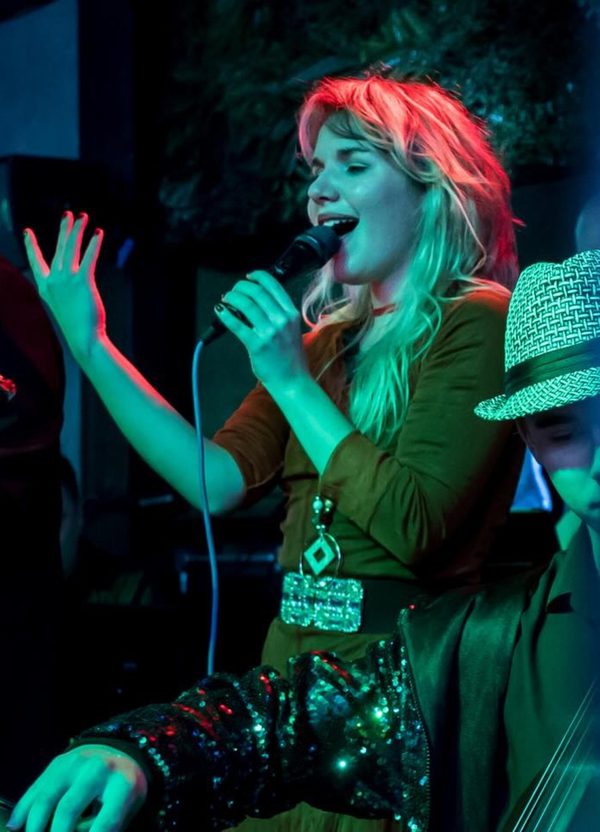 A band performing on stage under blue lighting, featuring a blonde female singer in the foreground, with a neon sign glowing behind them.