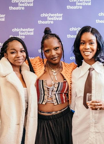 Three women smile at the camera. Behind them is a blue background with the Chichester Festival Theatre logo.