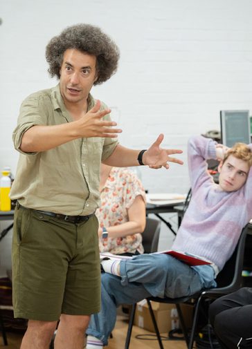 A man in a pale green shirt and khaki shorts gesticulates while talking in a rehearsal room. On the right, a young man in a purple jumper is listening intently.