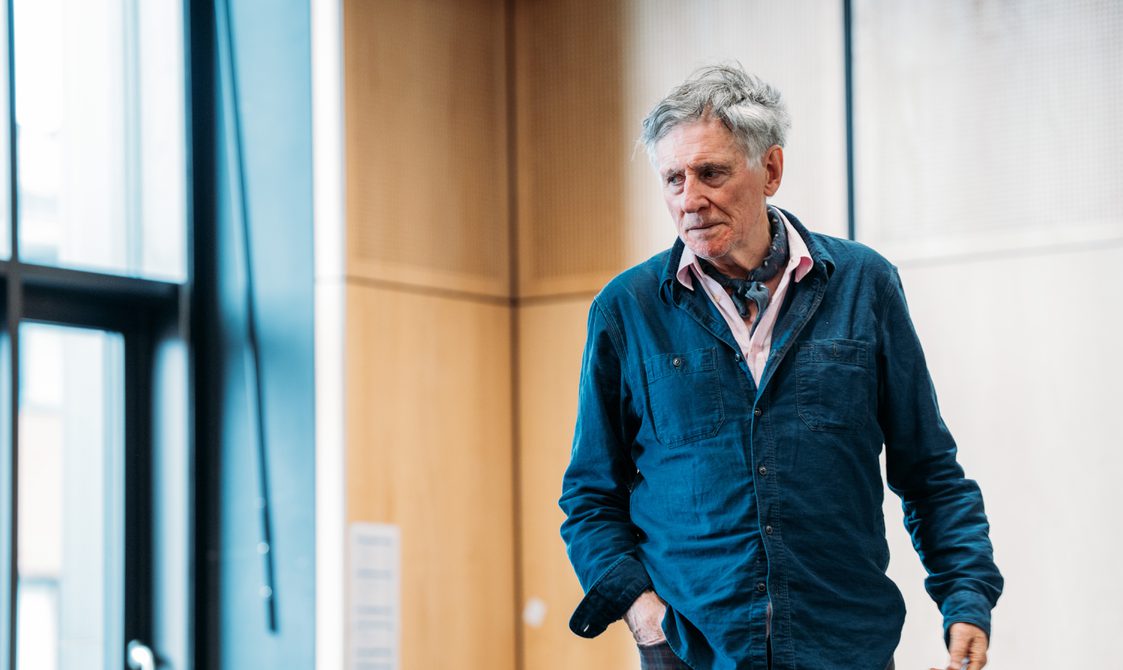 An actor in a blue overshirt stands in a rehearsal room, he holds a serious expression on his face.