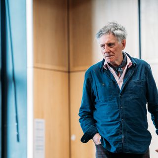 An actor in a blue overshirt stands in a rehearsal room, he holds a serious expression on his face.