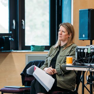 A sound desk is set up on the right hand side of this image to the left a woman sits on a chair with her legs crossed over one another holding a script.