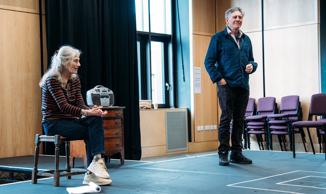 Two Actors stand on a stage in a rehearsal room, the woman on the left sits on a stool with her left leg cross over her right, she holds her hands together the man on the right stands with a wistful expression.