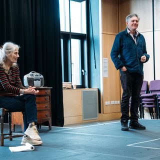 Two Actors stand on a stage in a rehearsal room, the woman on the left sits on a stool with her left leg cross over her right, she holds her hands together the man on the right stands with a wistful expression.