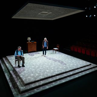 A man and woman are on a minimal set. The floor is a large carpeted square, above them is a smaller square suspended from the ceiling. She stands, facing towards him, as he sits in a chair with a book in his hands.