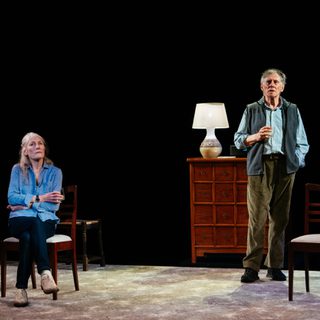 An older man is stood by a chest of drawers, he looks out front ahead of him as he holds a drink. He wears corduroy trousers, a blue check shirt and a gilet. To his left, an older woman is sat in a chair with her arms folded, she has a contemplative expression. She is wearing a blue shirt and dark trousers.