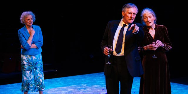 Three actors are stood on a minimal set. The floor is a large carpeted square, above them is a smaller square, glowing blue, suspended from the ceiling. At the front of the stage, a man and woman are stood close together looking out as the man points to something in the distance. Behind them, another woman looks on.